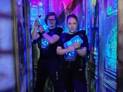 Two women in neon-lit laser tag arena wearing vests and holding glowing blue laser guns, posing for the camera in a dark, colorful setting.